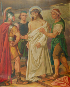 Ein Gemälde, das Jesus mit Dornenkrone und leuchtendem Heiligenschein zeigt, der von drei römischen Soldaten seines Gewandes entkleidet wird; Jesus steht ruhig da, während die Soldaten in antiker Kleidung ernst und konzentriert schauen. (Text mit KI erstellt)