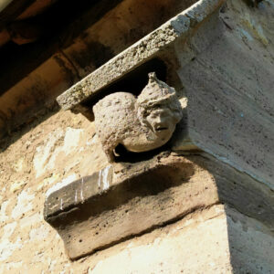 Eine verwitterte Steinskulptur eines Gesichts mit einem spitzen Hut oder Helm, Teil eines architektonischen Elements an der Ecke eines alten Gebäudes, das vom Sonnenlicht beleuchtet wird. (Text mit KI erstellt)