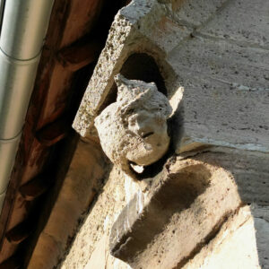 Eine verwitterte Steinskulptur eines Gesichts mit einem spitzen Hut ist in eine Außenwand eingelassen, die teilweise vom Sonnenlicht beleuchtet wird, in der Nähe einer Metalldachrinne und eines Holzdachs. (Text mit KI erstellt)