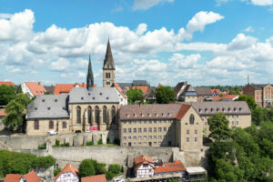 Luftaufnahme einer historischen europäischen Stadt mit steinernen Gebäuden, roten Ziegeldächern und Kirchtürmen unter einem blauen Himmel mit vereinzelten Wolken. Üppige grüne Bäume umgeben die Gebäude. (Text mit KI erstellt)