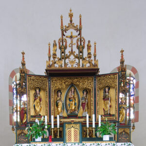 Ein verzierter Kirchenaltar mit goldenen Verzierungen, religiösen Statuen und offenen Tafeln mit Heiligenbildern. Weiße Kerzen, grüne Pflanzen und ein dekoratives Tuch sind vor dem Altar aufgestellt. Dahinter sind Buntglasfenster zu sehen. (Text mit KI erstellt)