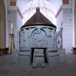 Ein steinernes Taufbecken mit geschnitzten religiösen Figuren und einem spitzen Metalldeckel, flankiert von skulptierten Löwen, steht in einem Kirchenraum mit gewölbten Decken und Bänken. (Text mit KI erstellt)