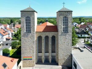 Eine Steinkirche mit zwei hohen quadratischen Türmen, Bogenfenstern und einem roten Ziegeldach steht inmitten eines Wohngebiets. Neben dem Eingang hängt ein Regenbogenbanner. Bäume und Häuser umgeben das Gebäude unter einem blauen Himmel. (Text mit KI erstellt)
