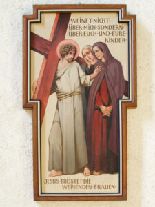 Eine gemalte religiöse Plakette zeigt Jesus, der ein Kreuz trägt und zwei weinende Frauen tröstet. Der deutsche Text lautet: Weinet nicht über mich, sondern über euch und eure Kinder. Jesus tröstet die weinenden Frauen. (Text mit KI erstellt)