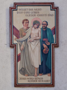 Religiöses Gemälde von Jesus mit Dornenkrone und Heiligenschein, flankiert von zwei Männern; einer hält einen Krug. Der deutsche Text darüber und darunter bezieht sich auf die Entkleidung Jesu. Das Bild ist in Form eines Kreuzes gerahmt. (Text mit KI erstellt)