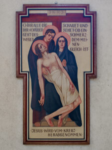 Ein religiöses Gemälde zeigt Jesus, der von zwei Trauernden vom Kreuz genommen wird. Ein deutscher Text umgibt die Figuren. Das Bild ist von einer dekorativen Bordüre umrahmt und an einer hellen Wand angebracht. (Text mit KI erstellt)