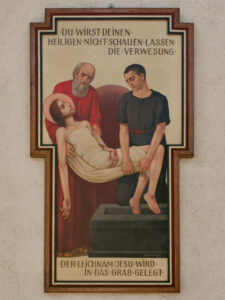 Ein religiöses Gemälde zeigt zwei Männer, die den leblosen, in ein Tuch gewickelten Körper Jesu in ein Grab legen. Ein deutscher Text darüber und darunter beschreibt die Szene. Das Kunstwerk ist in einem hölzernen, kreuzförmigen Rahmen an einer Wand angebracht. (Text mit KI erstellt)