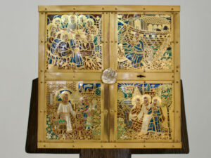 Ein goldenes religiöses Altarbild mit vier geprägten Tafeln, auf denen biblische Szenen, komplizierte Muster und Figuren dargestellt sind, montiert auf einem Holzständer vor einem schlichten Hintergrund. (Text mit KI erstellt)