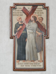 Ein religiöses Gemälde zeigt Jesus, der ein Holzkreuz trägt, flankiert von zwei Männern. Ein deutscher Text umgibt die Szene, und das Kunstwerk ist in Form eines Kreuzes gerahmt und an einer strukturierten Wand angebracht. (Text mit KI erstellt)