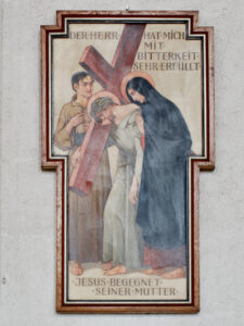 Eine gemalte Darstellung von Jesus, der das Kreuz trägt und seiner Mutter Maria begegnet, mit einem Umstehenden hinter ihnen. Der deutsche Text oben und unten beschreibt die Szene, in der Jesus seiner Mutter begegnet. Das Bild ist in Holz gerahmt. (Text mit KI erstellt)