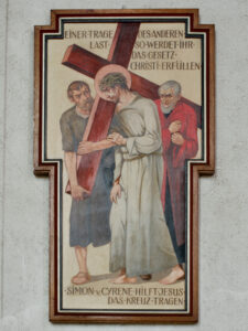 Ein Gemälde zeigt Simon von Cyrene, der Jesus beim Tragen des Kreuzes hilft, während ein römischer Soldat sie führt. Der deutsche Text darüber und darunter beschreibt die biblische Szene. Das Kunstwerk ist in einer kreuzförmigen Umrahmung an einer Wand angebracht. (Text mit KI erstellt)