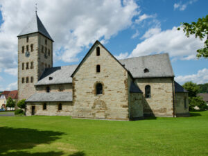 Eine historische Steinkirche mit einem hohen Glockenturm und einem Schieferdach steht auf einem grünen Rasen unter einem blauen Himmel mit vereinzelten Wolken. Im Hintergrund sind Bäume und Gebäude zu sehen. (Text mit KI erstellt)
