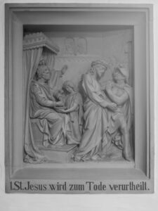 Eine Reliefskulptur stellt Jesus dar, der von einem sitzenden Beamten zum Tode verurteilt wird, wobei mehrere Figuren anwesend sind. Der deutsche Text darunter lautet: 1. St. Jesus wird zum Tode verurteilt. (Text mit KI erstellt)