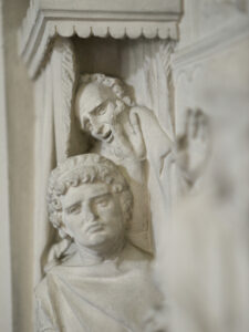 Nahaufnahme einer Steinreliefskulptur mit zwei Figuren; die vordere Figur hat lockiges Haar und einen ruhigen Gesichtsausdruck, während die hintere Figur ein dramatisches Gesicht hat und eine Hand hebt, als würde sie flüstern oder gestikulieren. (Text mit KI erstellt)