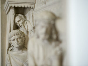 Nahaufnahme eines Steinreliefs mit drei menschlichen Figuren, zwei im Hintergrund mit ausdrucksstarken Gesichtern und einer unscharfen Figur im Vordergrund, die alle sehr detailliert geschnitzt sind. (Text mit KI erstellt)
