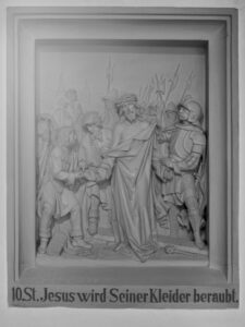 Eine Reliefskulptur zeigt Jesus, der von mehreren Männern entkleidet wird, umgeben von römischen Soldaten mit Speeren. Die Inschrift darunter lautet: 10. St. Jesus wird Seiner Kleider beraubt. (Text mit KI erstellt)