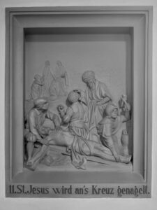 Eine Reliefskulptur, die den ans Kreuz genagelten Jesus darstellt, der von mehreren trauernden Figuren umgeben ist. Die Inschrift darunter lautet: 11. St. Jesus wird ans Kreuz genagelt. (Text mit KI erstellt)
