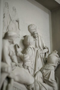 Ein detailliertes Steinrelief zeigt mehrere Figuren in historischer Kleidung, darunter Männer in Gewändern und Turbanen, die sich mit aufmerksamer Miene unterhalten. Im Hintergrund sind schwache Umrisse weiterer Figuren zu erkennen. (Text mit KI erstellt)