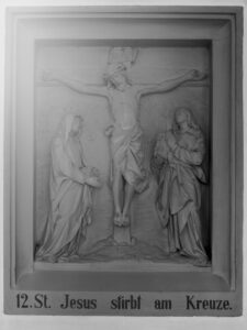 Eine Reliefskulptur, die den gekreuzigten Jesus mit zwei trauernden Figuren neben ihm zeigt. Die Inschrift darunter lautet: 12. St. Jesus stirbt am Kreuze. (Text mit KI erstellt)