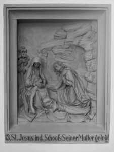 Ein Steinrelief zeigt Jesus, der nach der Kreuzigung in den Schoß seiner Mutter gelegt wird, mit Trauernden an ihrer Seite. Der deutsche Text darunter lautet 13. St. Jesus ind. Schoofs Seiner Mutter gelegi. (Text mit KI erstellt)