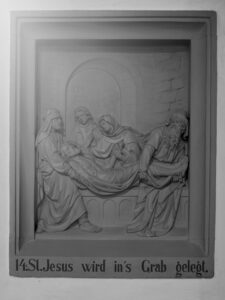 Ein Steinrelief stellt vier Trauernde dar, die Jesus in ein Grab legen. Darunter steht in deutscher Sprache: 14: St. Jesus wird ins Grab gelegt (14: Jesus wird ins Grab gelegt). Die Szene wird von einer rechteckigen Umrandung eingerahmt. (Text mit KI erstellt)