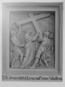 Ein Steinrelief zeigt Jesus, der ein Kreuz auf der Schulter trägt, unterstützt und umgeben von mehreren Figuren. Darunter steht ein deutscher Text: 2. St. Jesus nimmt d. Kreuz auf Seine Schultern. (Text mit KI erstellt)