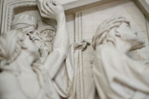 Ein detailliertes Steinrelief zeigt mehrere Figuren in antiker Kleidung, die nach rechts blicken, wobei eine zentrale Figur auf etwas zeigt und andere aufmerksam zusehen, was auf eine dramatische oder historische Szene hindeutet. (Text mit KI erstellt)