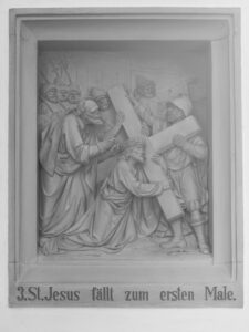 Reliefskulptur, die Jesus darstellt, der unter der Last des Kreuzes fällt, umgeben von Soldaten und Anhängern. Die Inschrift darunter lautet 3. St. Jesus fällt zum ersten Male, was auf die dritte Station des Kreuzweges hinweist. (Text mit KI erstellt)
