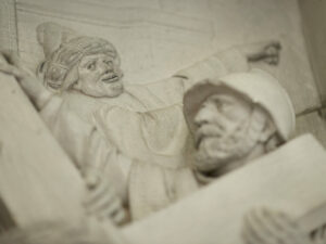 Nahaufnahme einer Steinreliefskulptur, die zwei Figuren zeigt: eine mit Turban und Gewand, die mit ausgestrecktem Arm gestikuliert, und eine andere mit Helm, die in die gleiche Richtung blickt; beide scheinen in Aktion zu sein. (Text mit KI erstellt)