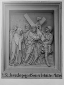 Ein Steinrelief zeigt Jesus, der ein Kreuz trägt und seiner trauernden Mutter begegnet, umringt von römischen Soldaten und Schaulustigen. Der deutsche Text darunter lautet: 4. St. Jesus begegnet seiner betrübten Mutter. (Text mit KI erstellt)