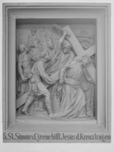 Ein plastisches Relief zeigt Simon von Cyrene, der Jesus beim Tragen des Kreuzes hilft, umgeben von Soldaten und Schaulustigen. Die Inschrift darunter lautet: "5. St. Simon v. Cyrene hilft Jesus d. Kreuz tragen.". (Text mit KI erstellt)