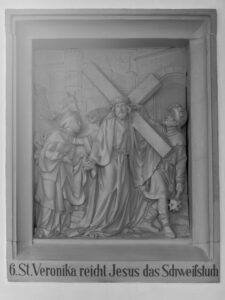 Eine Reliefskulptur, die die heilige Veronika darstellt, die Jesus ein Tuch reicht, während er das Kreuz trägt, umgeben von mehreren Figuren. Darunter befindet sich die deutsche Inschrift: 6. St. Veronika reicht Jesus das Schweißtuch. (Text mit KI erstellt)