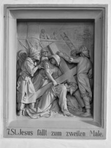 Ein detailliertes Steinrelief zeigt Jesus, der unter dem Kreuz fällt, umgeben von mehreren Figuren, die ihm Hilfe anbieten oder ihn anspornen. Darunter steht ein deutscher Text: 7. St. Jesus fällt zum zweiten Male. (Text mit KI erstellt)