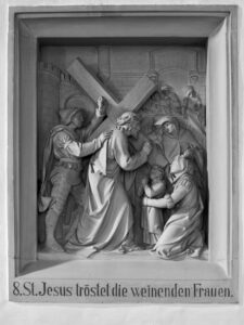 Ein Steinrelief zeigt Jesus, der ein Kreuz trägt und weinende Frauen und Kinder tröstet, während Soldaten in der Nähe stehen. Die Inschrift lautet: 8. St. Jesus tröstet die weinenden Frauen. (Text mit KI erstellt)