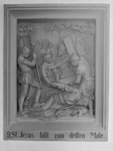 Ein Steinrelief stellt Jesus dar, der zum dritten Mal fällt, während er das Kreuz trägt, umgeben von römischen Soldaten und Schaulustigen. Die Inschrift darunter lautet: 9. St. Jesus fällt zum dritten Male. (Text mit KI erstellt)