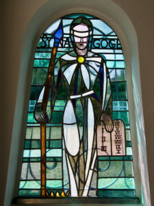 Glasfenster mit der Darstellung einer gewandeten Figur mit verbundenen Augen, die einen Stab und eine Steintafel mit den römischen Ziffern I-X in der Hand hält. Der Hintergrund ist in Blau- und Grüntönen gehalten, darüber steht das Wort SYNAGOGE. (Text mit KI erstellt)