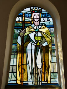 Glasfenster mit der Darstellung einer gekrönten Figur in weißem und goldenem Gewand, die einen Stab in der einen und einen Kelch in der anderen Hand hält, mit blauen und grünen geometrischen Hintergrundmustern. (Text mit KI erstellt)