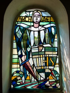 Glasfenster mit der Darstellung des Heiligen Michael, der mit Flügeln, Heiligenschein und Speer über einer Schlange steht. Der Name ST. MICHAEL ist oben in gelben und grünen Buchstaben geschrieben. Das Licht scheint durch das bunte Glas. (Text mit KI erstellt)