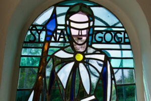 Glasfenster mit der Darstellung einer Figur, die einen blauen Stab hält und eine Augenbinde trägt. Das Wort SYNAGOGE ist im Hintergrund über den Schultern der Figur geschrieben. Draußen ist grünes Laub zu sehen. (Text mit KI erstellt)