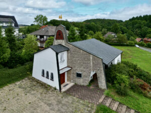 Umgeben von Bäumen und Häusern erhebt sich eine kleine, moderne Kirche mit weißem und steinernem Äußeren. Das Gebäude verfügt über einen Glockenturm mit einem goldenen Hahn auf der Spitze und einer Zeichnung eines Bootes an der Stirnwand. (Text mit KI erstellt)