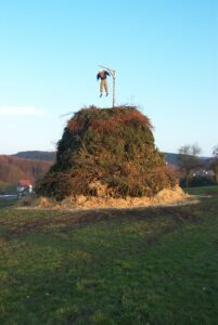 Ein großer Haufen aus Ästen und Gestrüpp steht auf einem Feld, an dessen Spitze ein Holzpfahl steht, an dem eine Vogelscheuchen-ähnliche Figur hängt. Die Szene spielt sich vor einer Kulisse aus Bäumen und Hügeln unter einem klaren Himmel ab. (Text mit KI erstellt)
