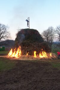 Auf einem offenen Feld brennt ein großes Lagerfeuer aus Ästen, an dessen Spitze eine menschenähnliche Figur oder ein Bildnis an einer Stange hängt. Bäume und Gras umgeben das Gebiet, und eine Person steht in der Nähe. (Text mit KI erstellt)