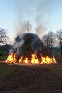 Ein großes Lagerfeuer brennt mit Flammen und aufsteigendem Rauch im Freien auf einer Wiese mit kahlen Bäumen im Hintergrund. Eine menschenähnliche Figur sitzt oben auf dem Haufen. Eine Person steht in der Nähe und hütet das Feuer. (Text mit KI erstellt)