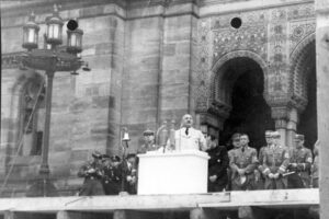Ein Mann spricht auf einem Podium vor einem großen, reich verzierten Gebäude, flankiert von uniformierten Offizieren und Soldaten. Mehrere Männer in Militäruniformen stehen in der Nähe, während eine Menschenmenge aufmerksam zuhört. (Text mit KI erstellt)