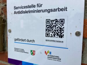 Ein Schild auf Deutsch an einer Wand mit der Aufschrift Servicestelle für Antidiskriminierungsarbeit enthält einen QR-Code, Logos und erwähnt die Integrationsagenturen NRW und das Ministerium für Arbeit, Gesundheit und Soziales. (Text mit KI erstellt)