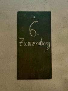 Eine dunkle rechteckige Kreidetafel mit der handgeschriebenen Zahl 6. und dem mit weißer Kreide geschriebenen Wort Zimmerberg, die an einer hellen Wand hängt. (Text mit KI erstellt)