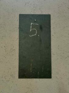 Eine rechteckige schwarze Schiefertafel ist an einer strukturierten grauen Wand angebracht. Die Zahl 5. ist mit weißer Kreide in der Nähe der oberen Mitte der Schiefertafel geschrieben. (Text mit KI erstellt)