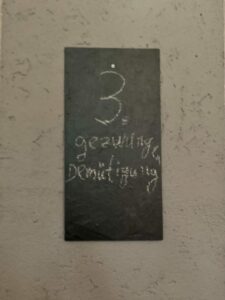 Ein dunkles rechteckiges Schild mit Kreideschrift hängt an einer strukturierten Wand. Oben steht eine große Zahl 3, darunter sind in Schreibschrift die Worte "Geswung" und "Demitsigung" zu lesen. (Text mit KI erstellt)
