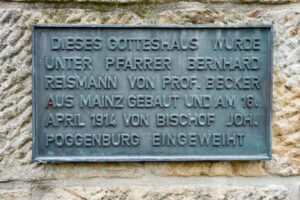 Eine Metalltafel an einer Steinwand mit deutschem Text besagt, dass die Kirche 1914 unter Pfarrer Bernhard Reismann und Prof. Becker aus Mainz erbaut und am 16. April 1914 von Bischof Joh. Poggenburg. (Text mit KI erstellt)