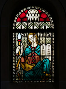 Glasfenster mit der Darstellung einer weiblichen Figur mit verbundenen Augen, die einen Stab hält, mit bunten Gewändern und einer Tafel mit römischen Ziffern; im Hintergrund sind architektonische und dekorative Motive zu sehen. (Text mit KI erstellt)
